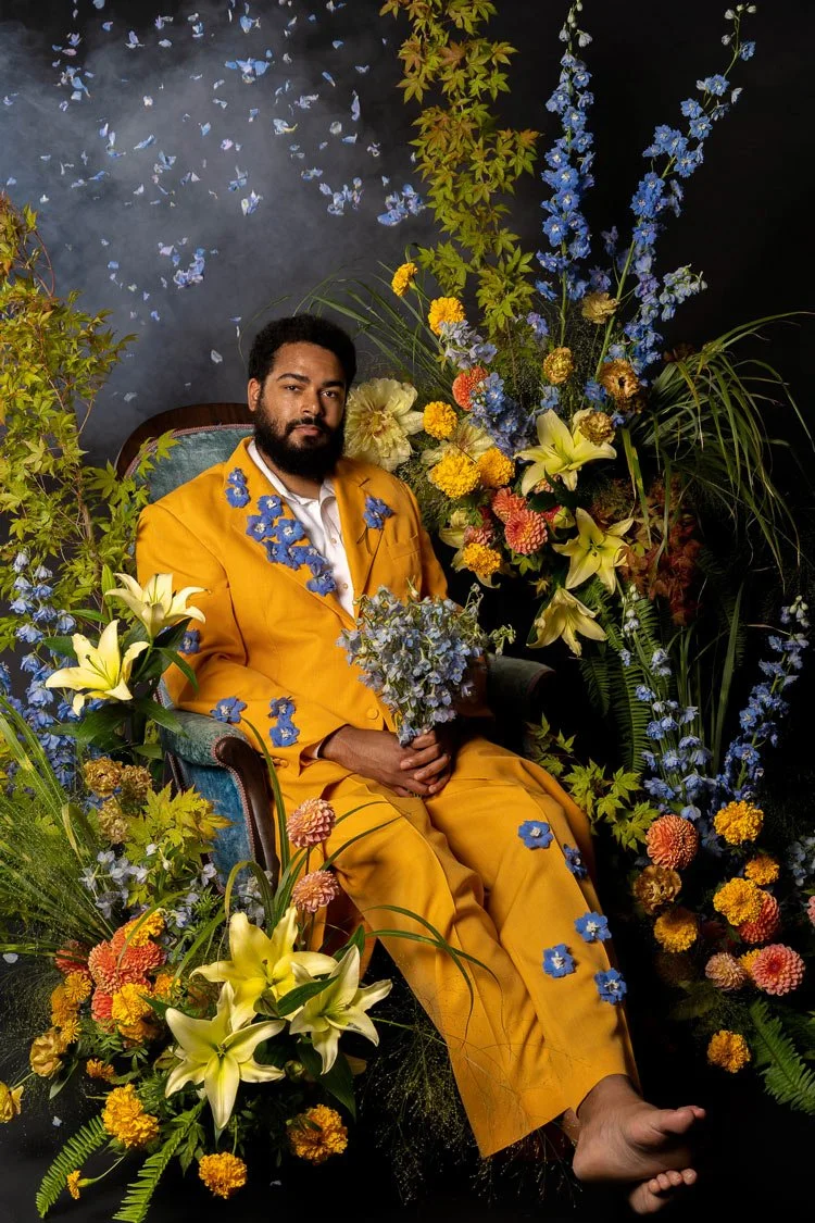 A fashion portrait featuring comedian Curtis Cook in a vibrant yellow suit with floral embellishments, surrounded by a lush and diverse arrangement of colorful flowers, set against a whimsical backdrop with light blue flower petals raining down.