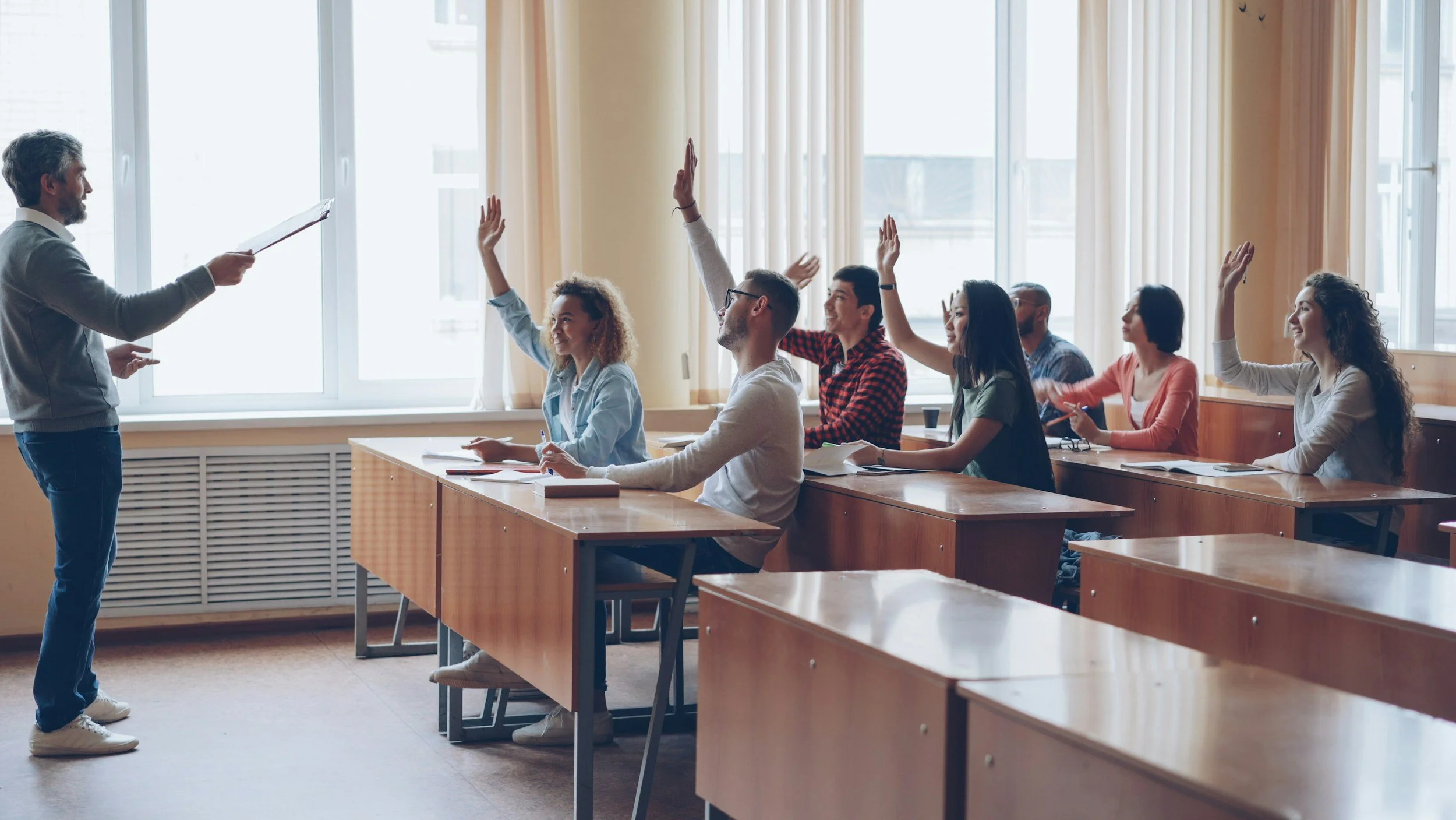 Teacher standing at the front of a classroom, holding a paper, with students raising their hands to answer questions.