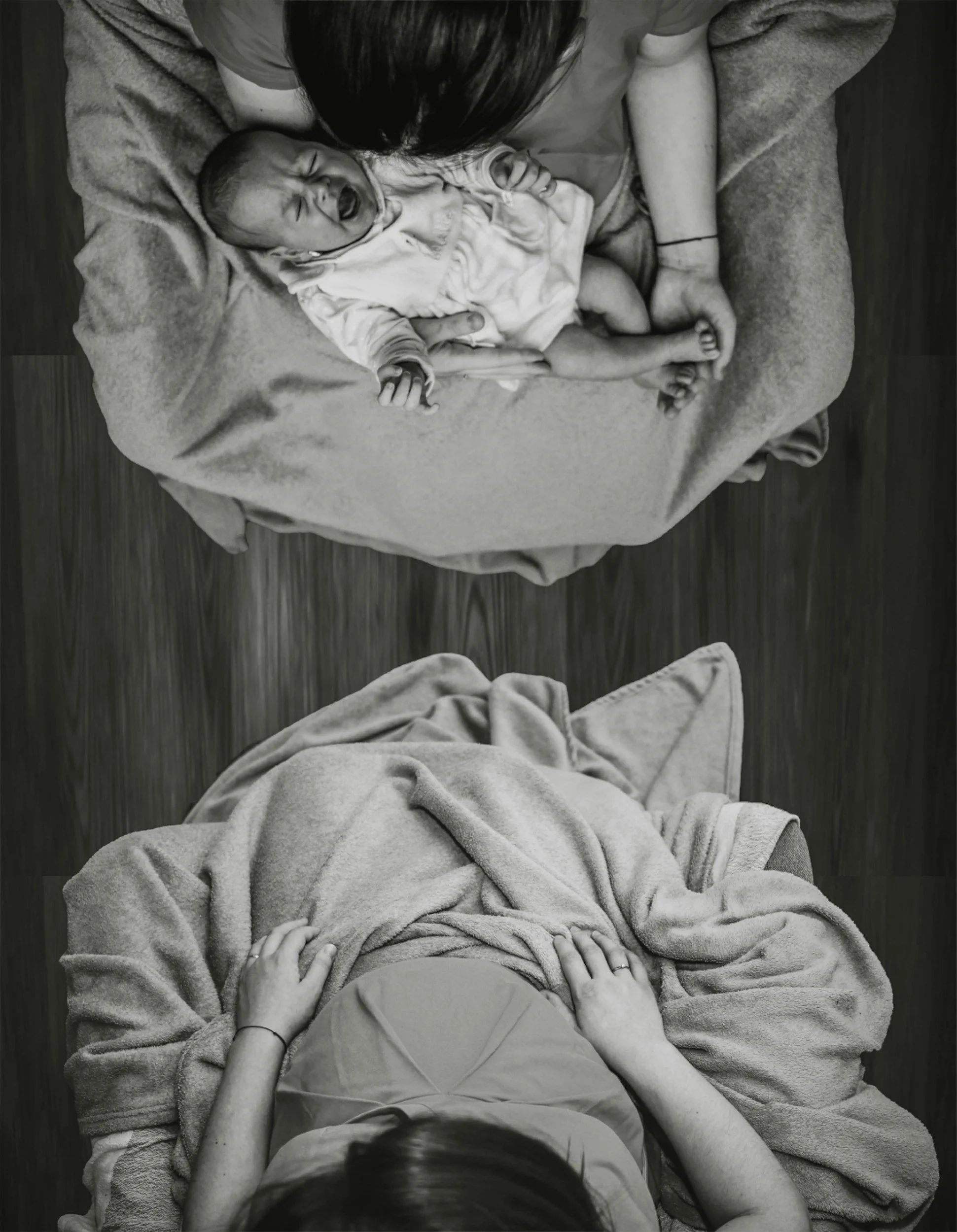 Black and white photo from above of a person holding a newborn baby, with the baby crying, on a bed with a blanket. Another person is lying below, their hands on their stomach, also on a bed.