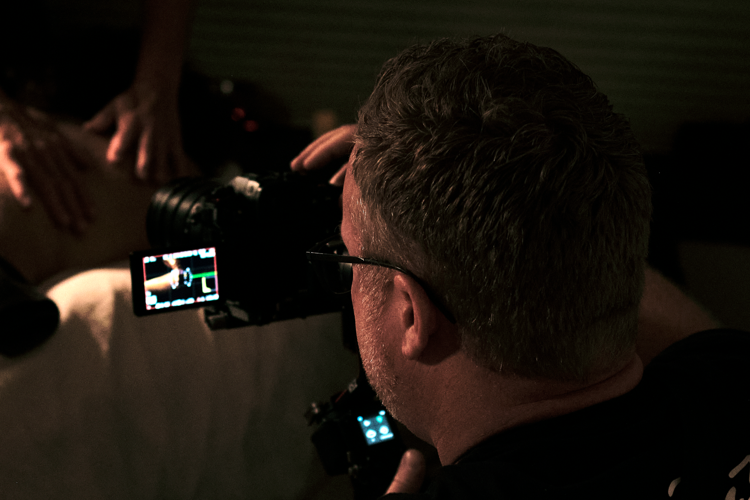 Darren is looking through a video camera's viewfinder, shooting content for a spa partner, in a dimly lit setting.