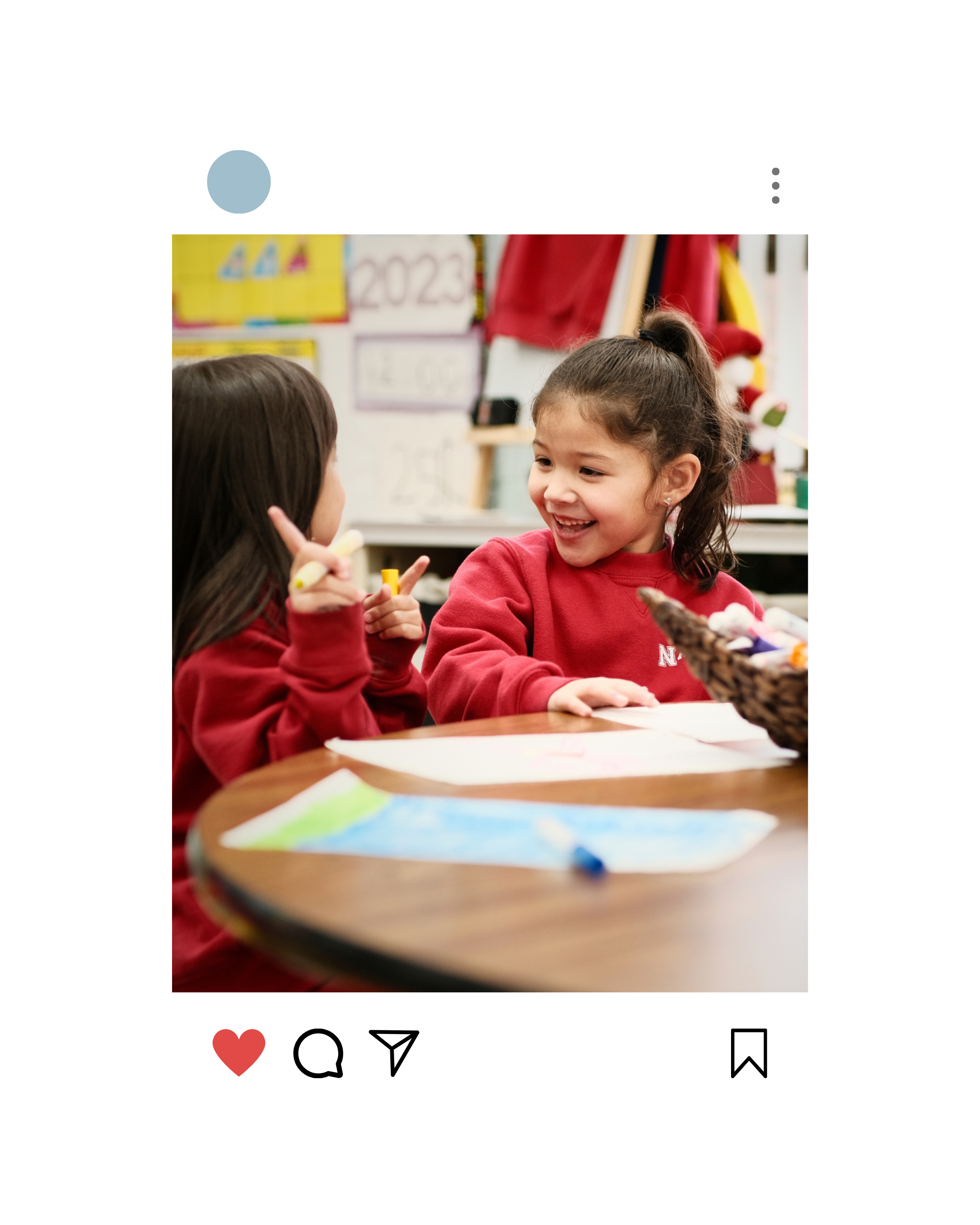 Social media graphic that showcases two young girls with brown hair, wearing red shirts, smiling and talking to each other at a classroom table, with art supplies and a colorful classroom in the background.