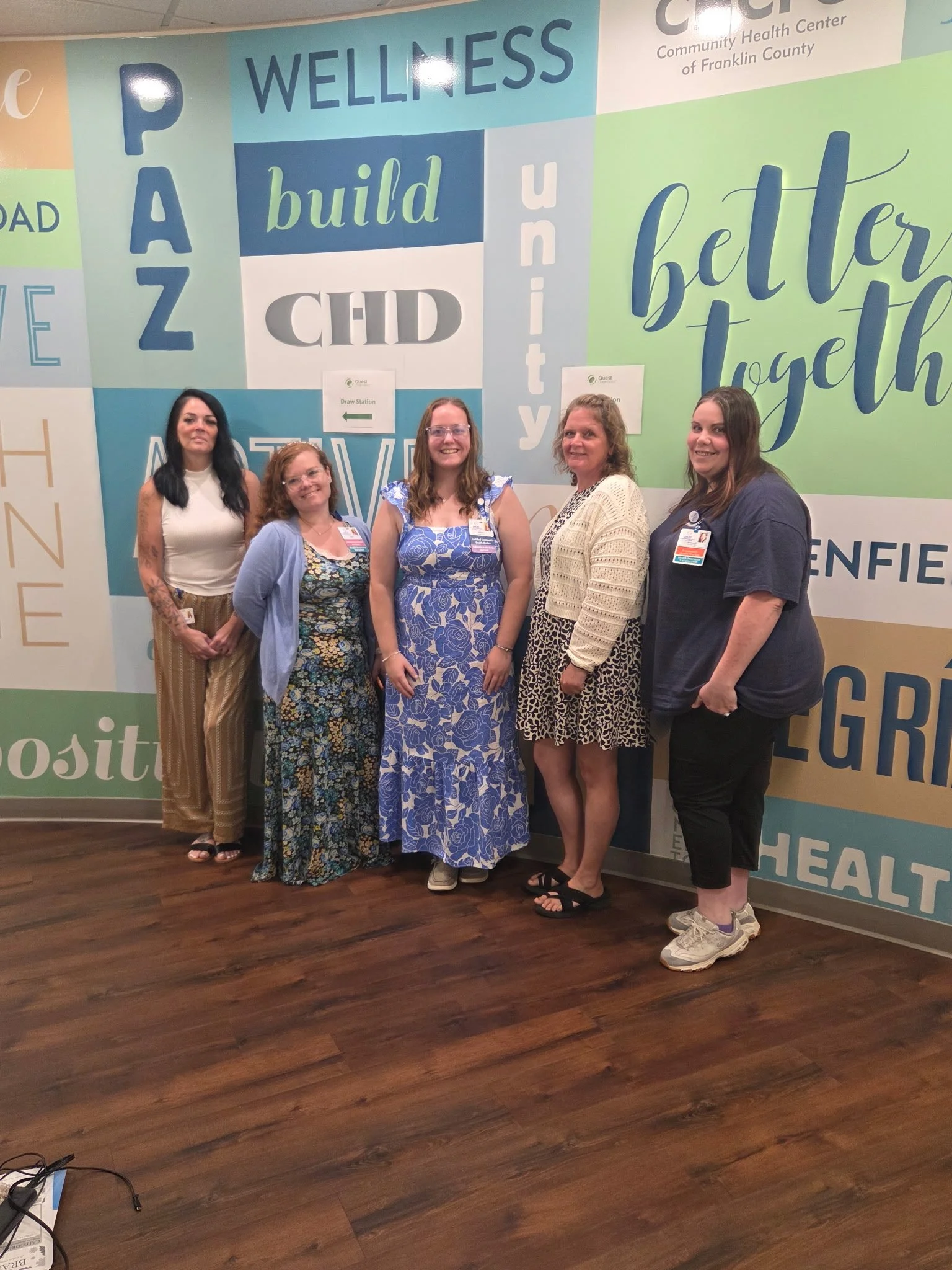 A photo of four women smiling in front of mural. These women compose our Community Health Worker Team in Orange and Greenfield.