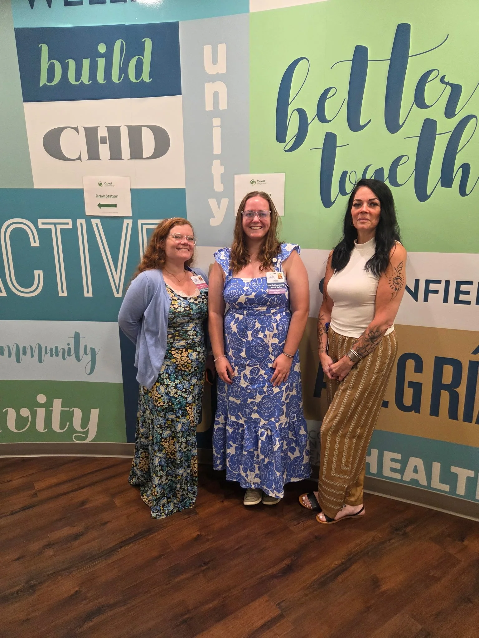 A photo of three smiling people in front of a mural. These folks are our Greenfield Community Health Worker Team.