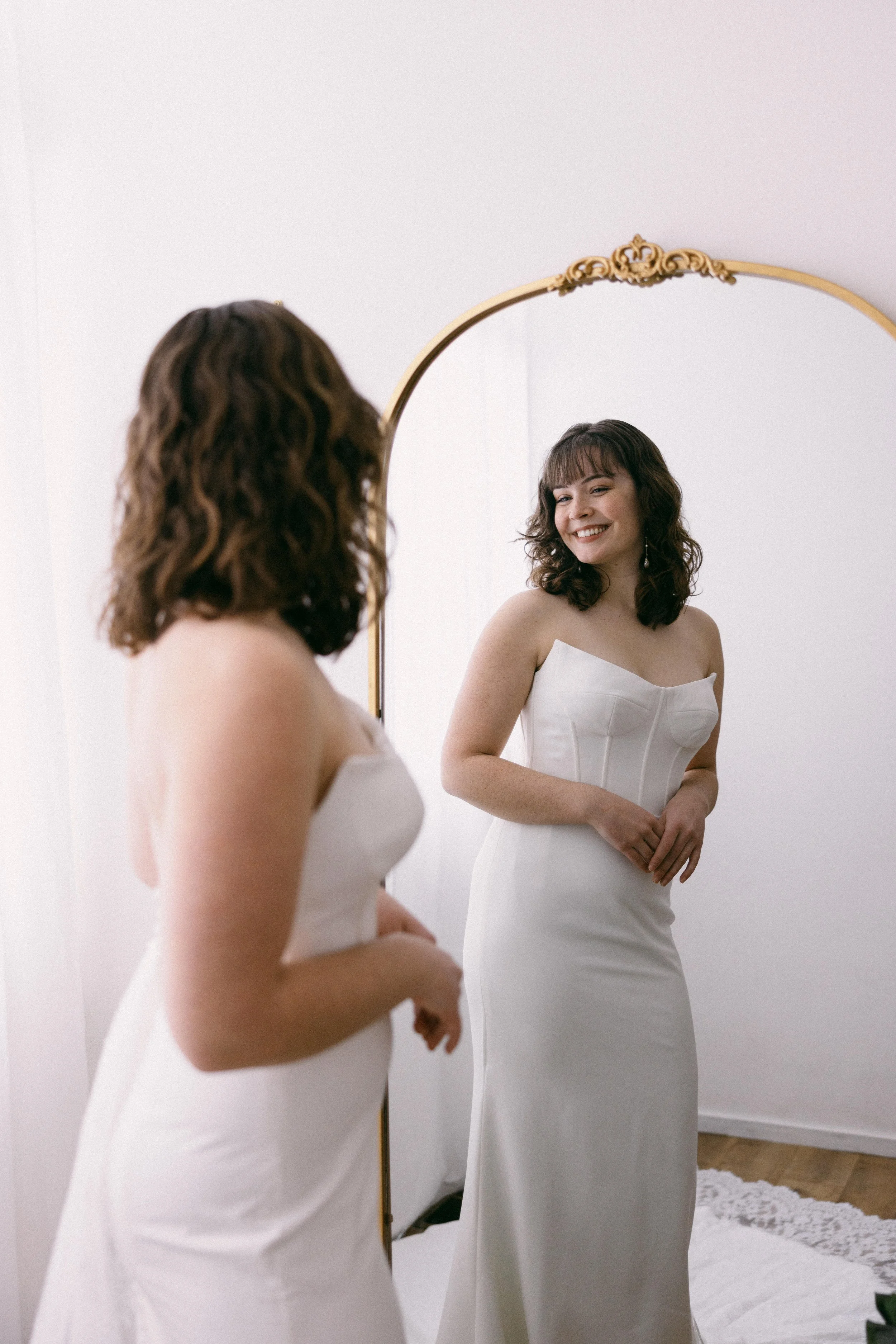bride in a fitted all white wedding dress looking fondly at herself in an ornate golden mirror