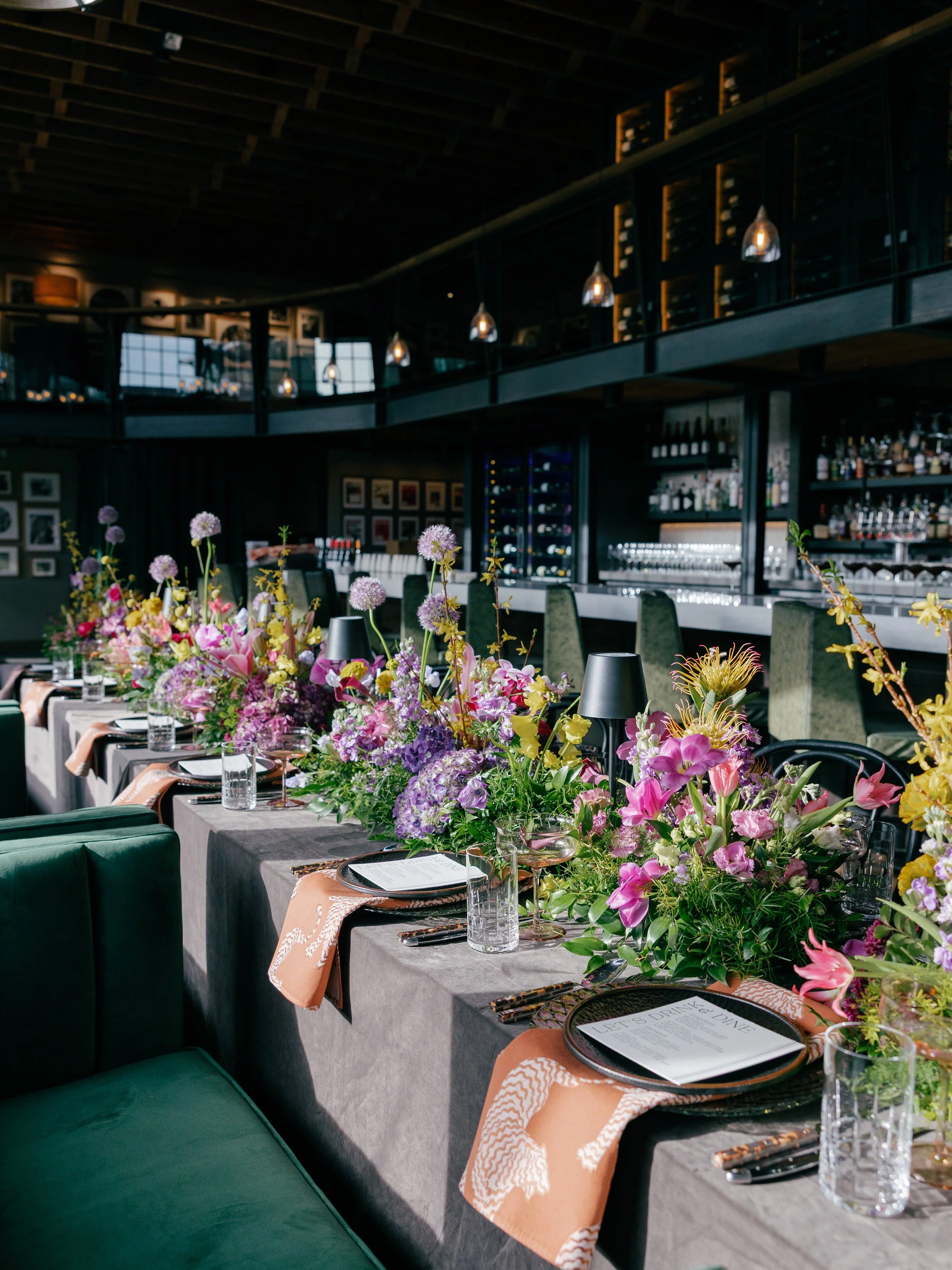 long-table-with-whimsical-bright-florals-down-the-center-in-a-high-ceiling-wine-cellar.jpg