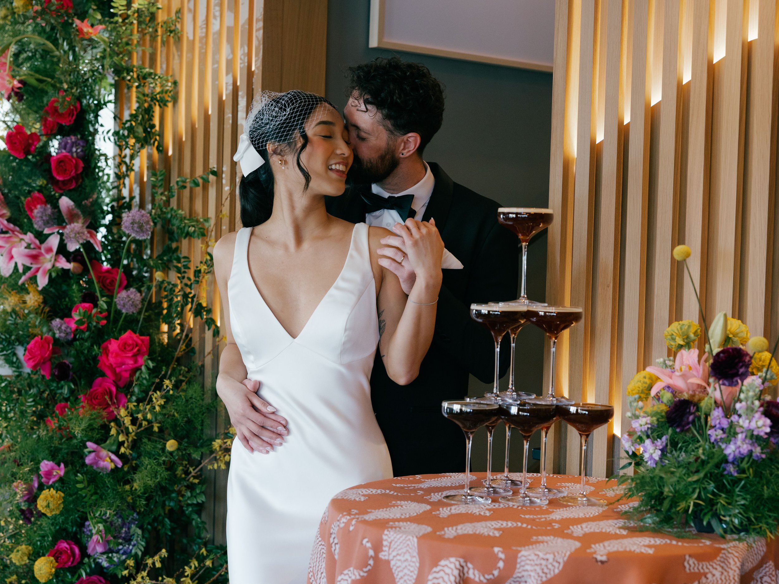 grooms-kissing-brides-neck-in-front-of-espresso-martini-tower.jpg
