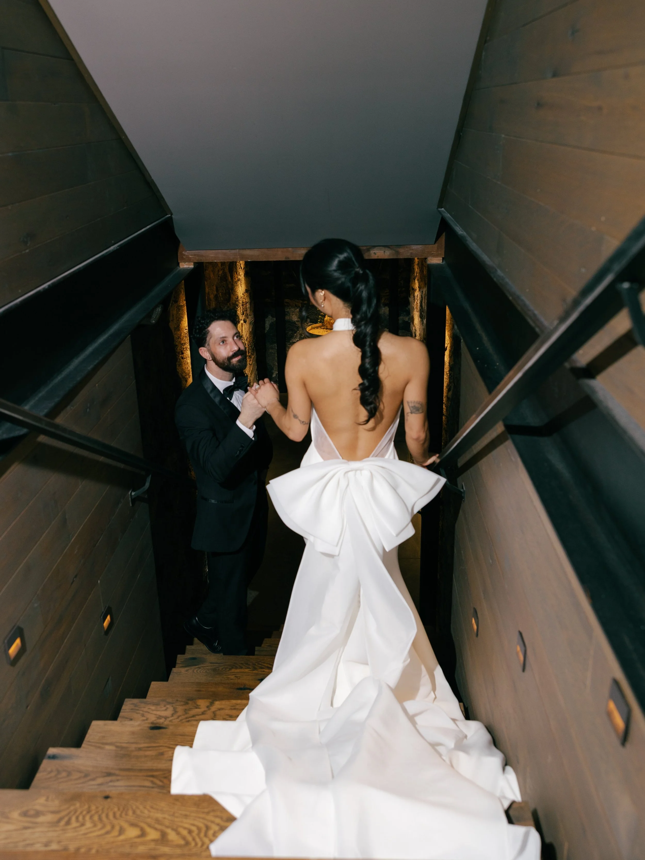 watching-bride-in-dress-with-big-bow-on-back-walk-down-stairs-into-moody-wine-cellar.jpg