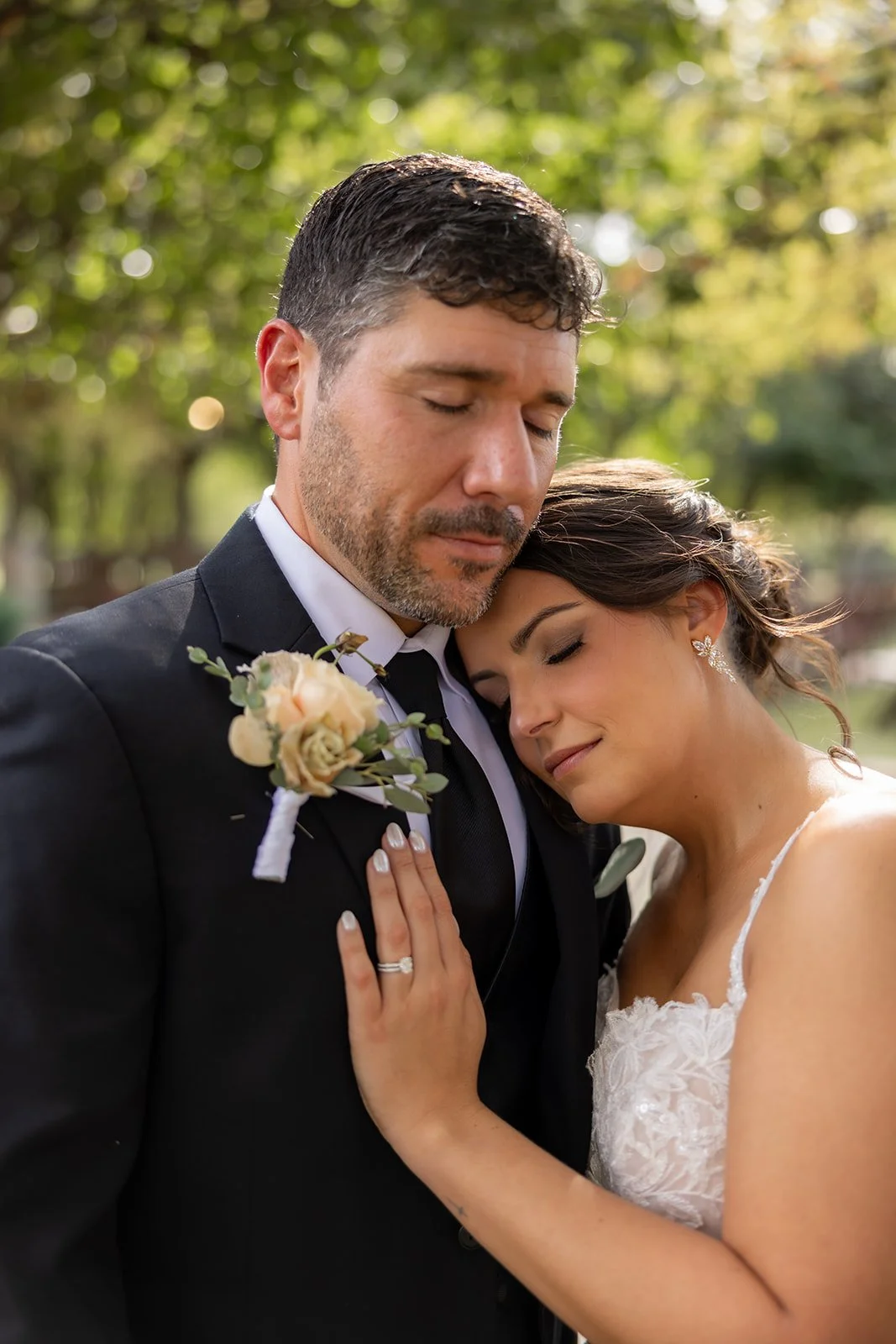 A bride and groom share an emotional embrace with eyes closed outdoors, surrounded by lush green trees.