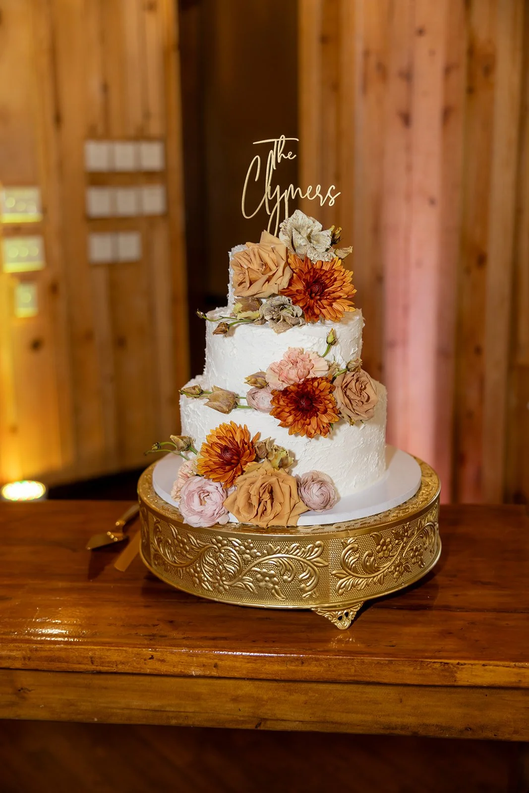 Two-tiered white wedding cake decorated with orange and pink flowers, topped with a gold cake topper that reads 'The Clymers,' on a gold ornate cake stand, on a wooden table with a wooden wall background.