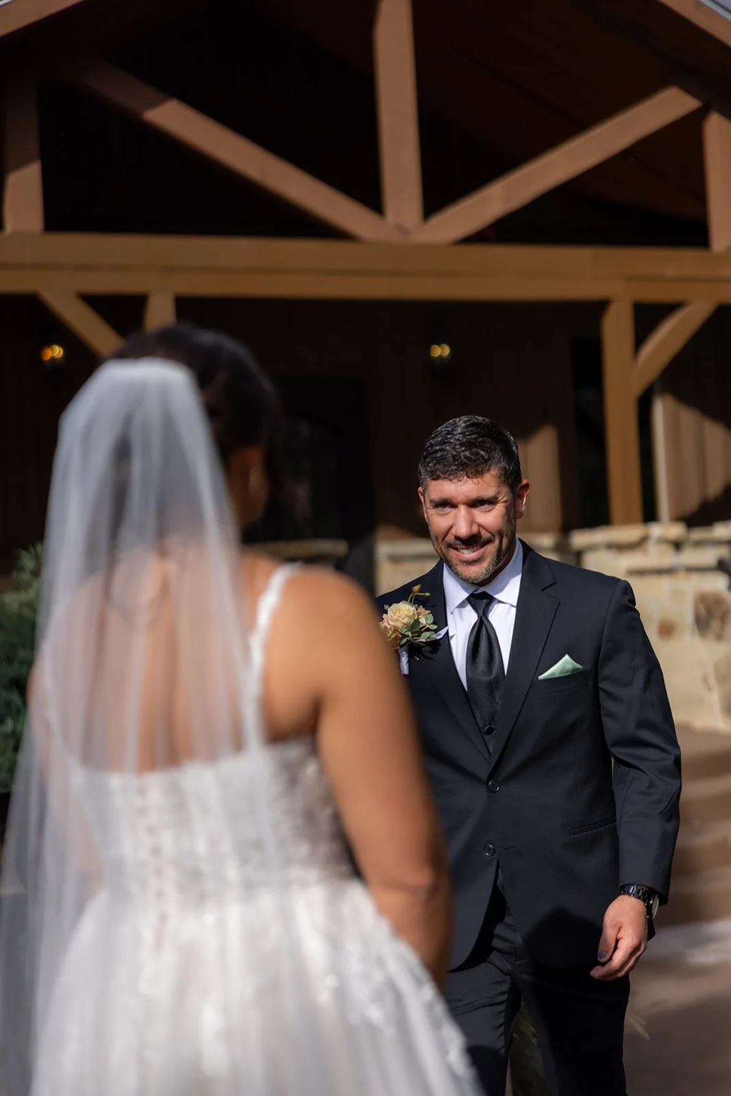 A groom smiling at a bride during a wedding ceremony in an indoor rustic venue with wooden beams.