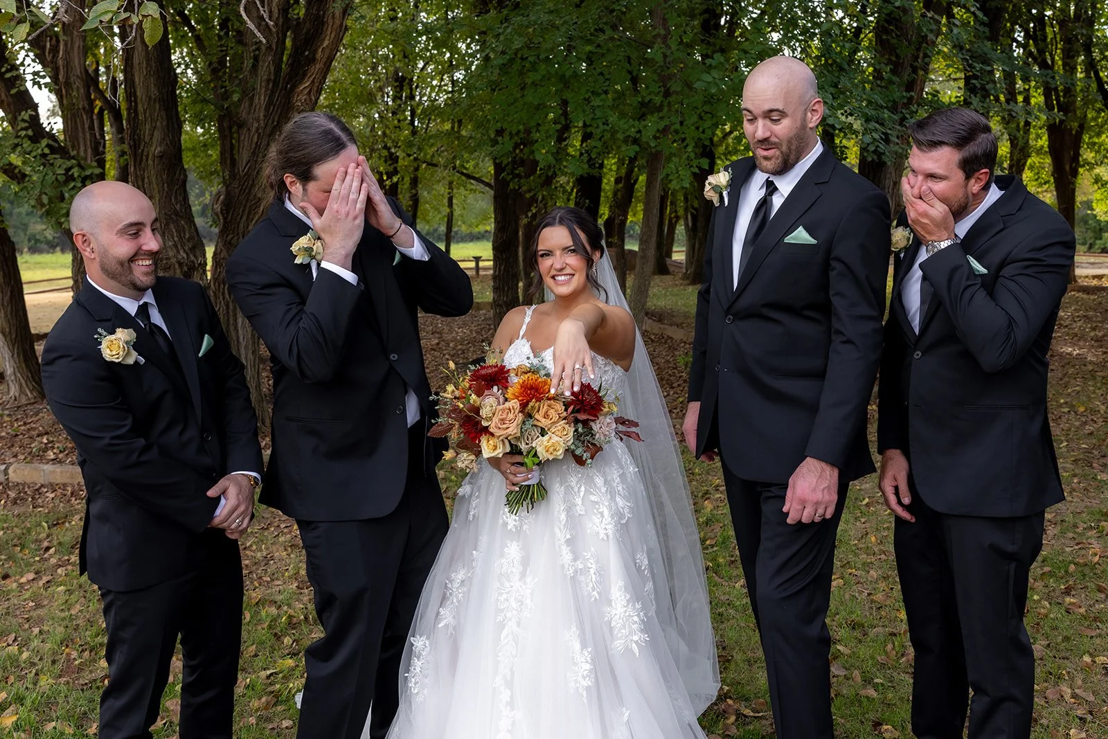 A bride in a white wedding dress and holding a bouquet of flowers stands among her groom and groomsmen in an outdoor park setting surrounded by trees, with some of the men reacting emotionally.