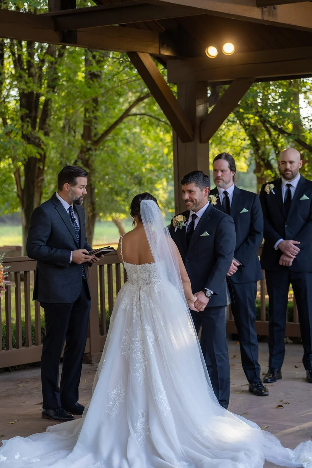Wedding ceremony with a bride and groom holding hands, standing under a wooden pavilion with officiant and groomsmen in suits, surrounded by green trees.