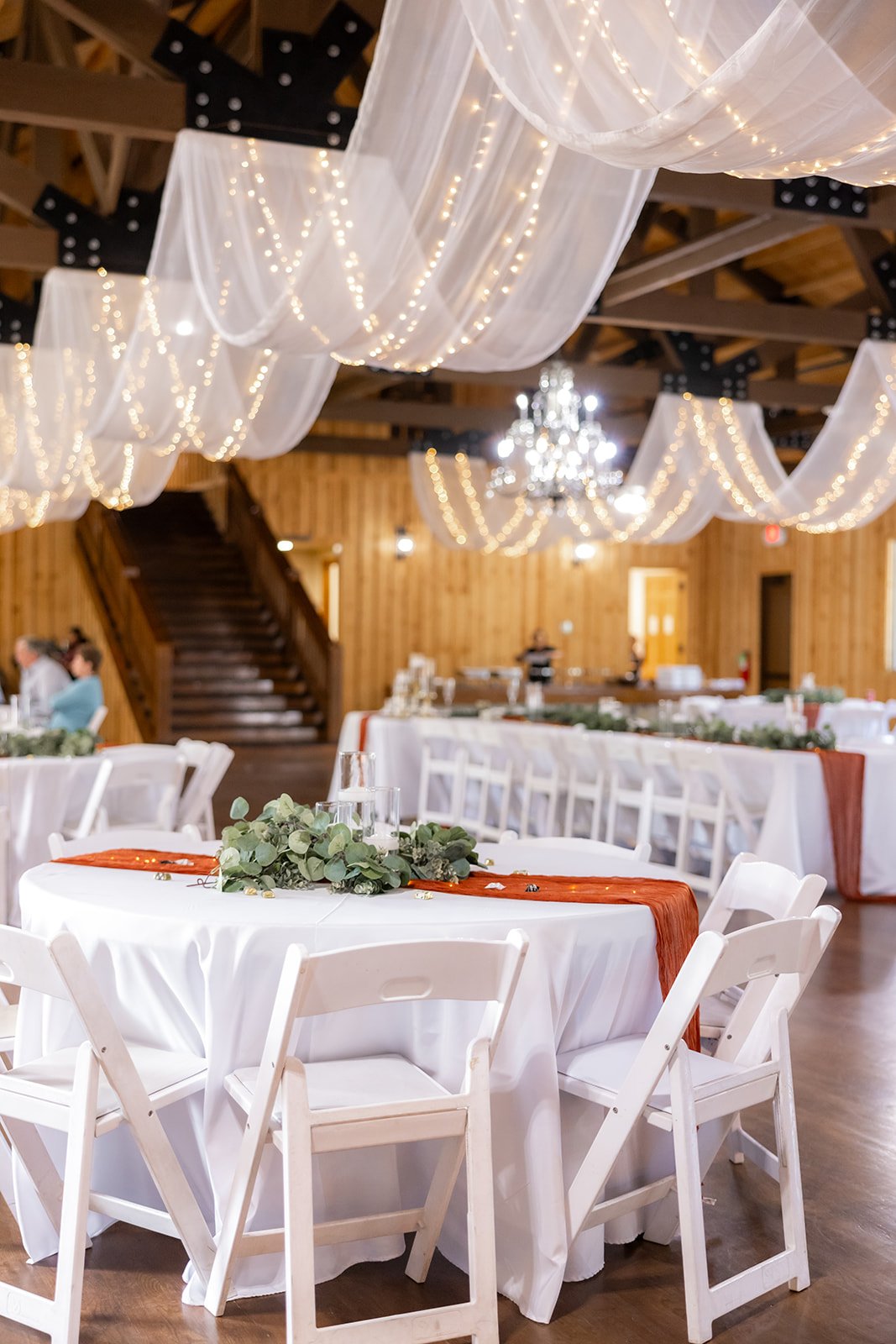 Wedding reception with decorated tables, white chairs, and hanging white drapes with string lights and a chandelier in a rustic wooden barn