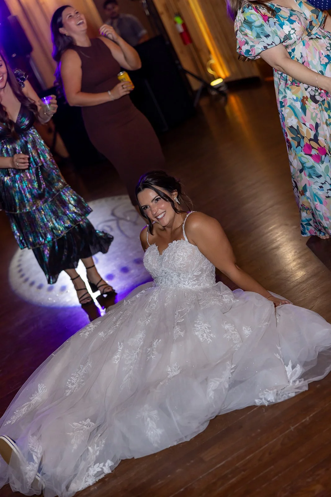 A woman in a white wedding dress sitting on the dance floor, smiling, with friends in the background at a celebration or wedding reception.