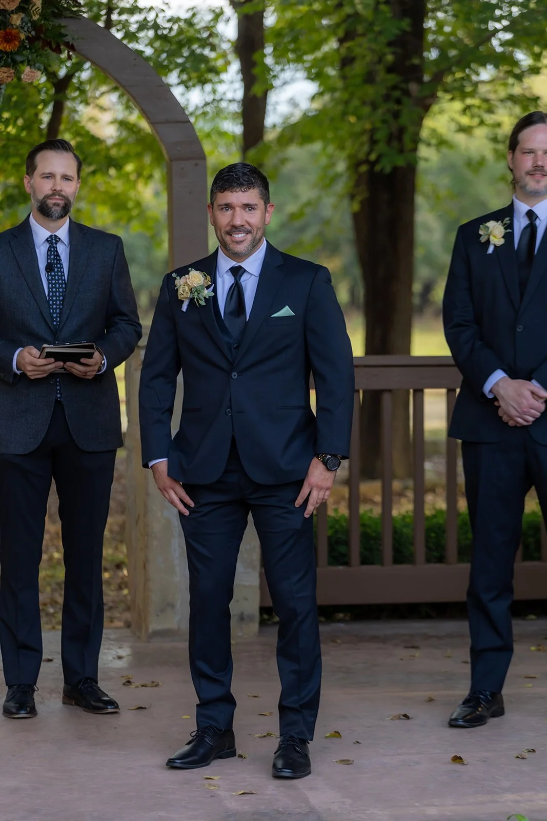 A groom in a navy suit with a boutonniere stands outdoors, flanked by two groomsmen in suits, with trees and a wooden arbor in the background.