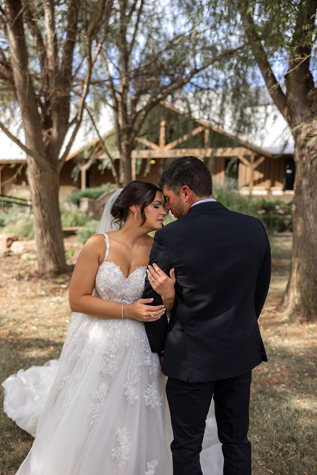 A bride and groom share an intimate moment outdoors in a wooded area, with the bride in a white lace wedding dress and the groom in a black tuxedo.