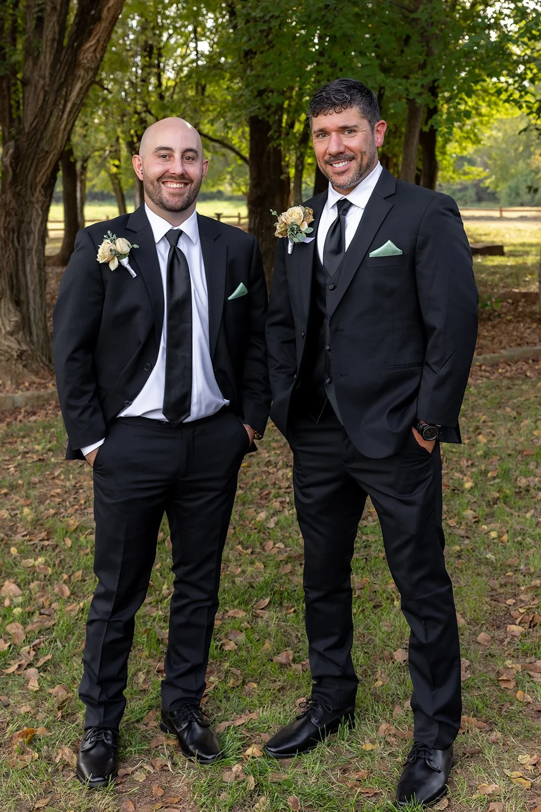 Two men in black suits with white shirts, black ties, and boutonnieres posing outdoors in a wooded area during daytime.