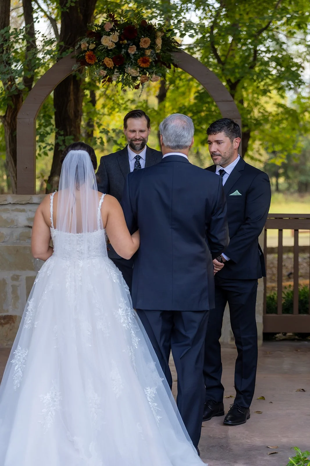 A bride and groom stand outdoors under an arch decorated with flowers, facing an officiant and another man, during a wedding ceremony in a natural setting with green trees in the background.