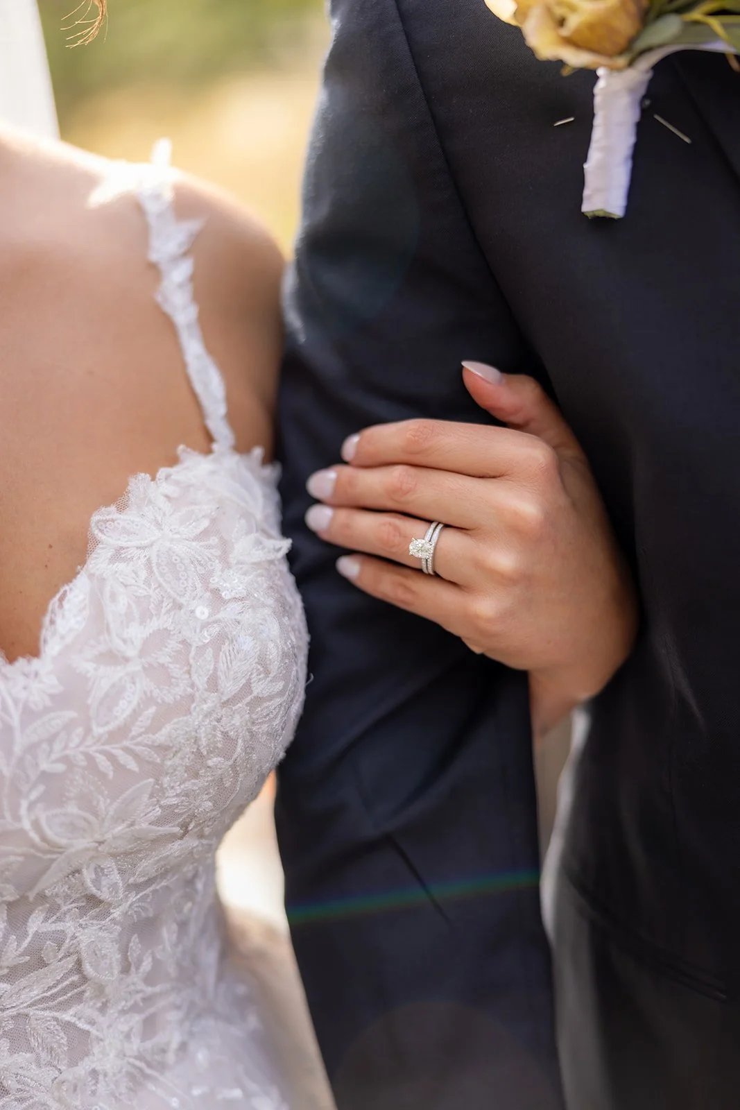 Close-up of a bride and groom on their wedding day, showing the bride's lace dress and wedding ring, and the groom's suit with a boutonniere, with sunlight in the background.