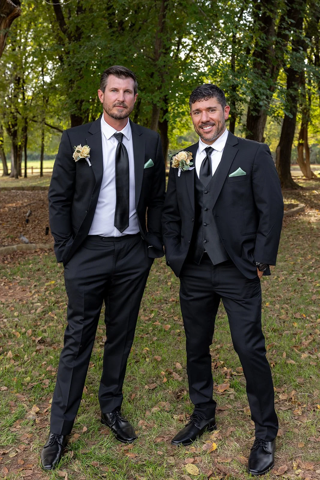 Two men in formal black suits, white shirts, and black ties standing outdoors in a wooded park during daytime. Both are wearing boutonnières and pocket squares, and have their hands in their pockets, smiling and posing for the photo.