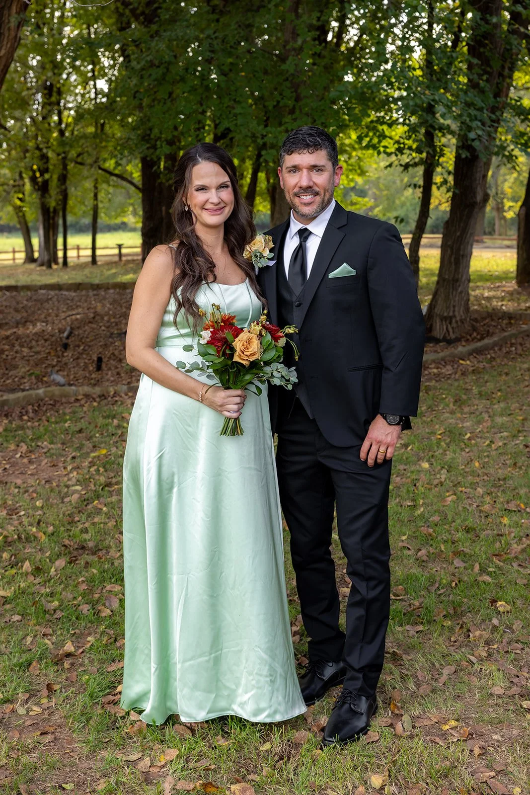 A bride and groom standing outdoors in a wooded area, dressed in wedding attire, smiling at the camera. The bride holds a bouquet of flowers, and the groom wears a black suit with a white shirt and tie.