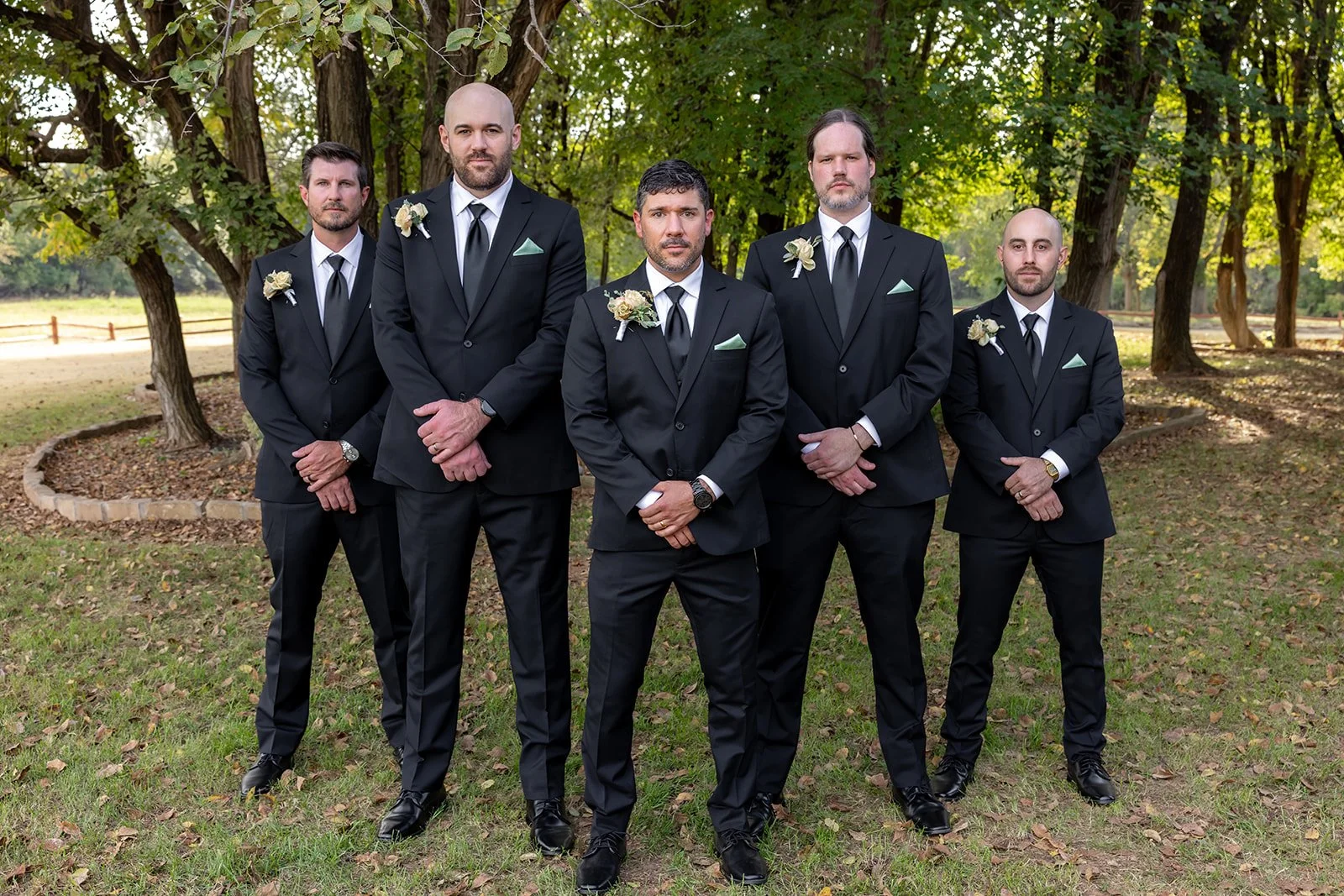 Five men dressed in black tuxedos with boutonnières, standing outdoors on a grassy area with trees in the background.