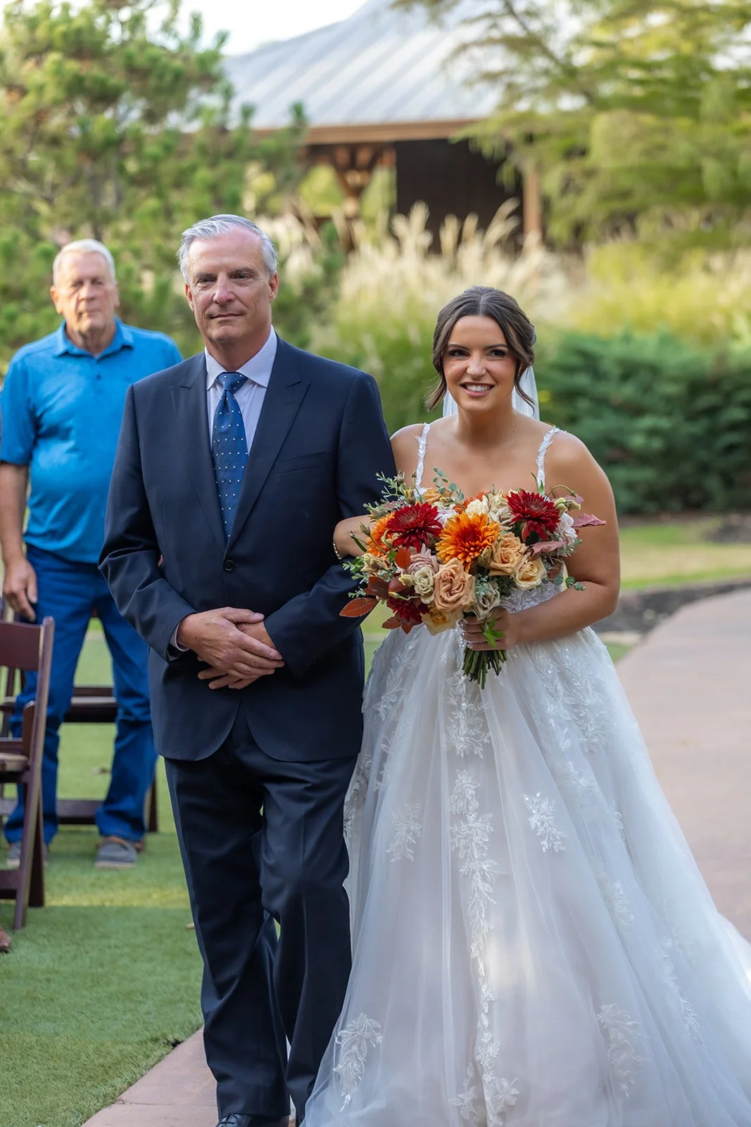 A bride in a white wedding gown holding a bouquet of orange, red, and beige flowers, walking down the aisle flanked by a man in a blue suit, with a man in a blue shirt in the background, outdoors in a garden setting.
