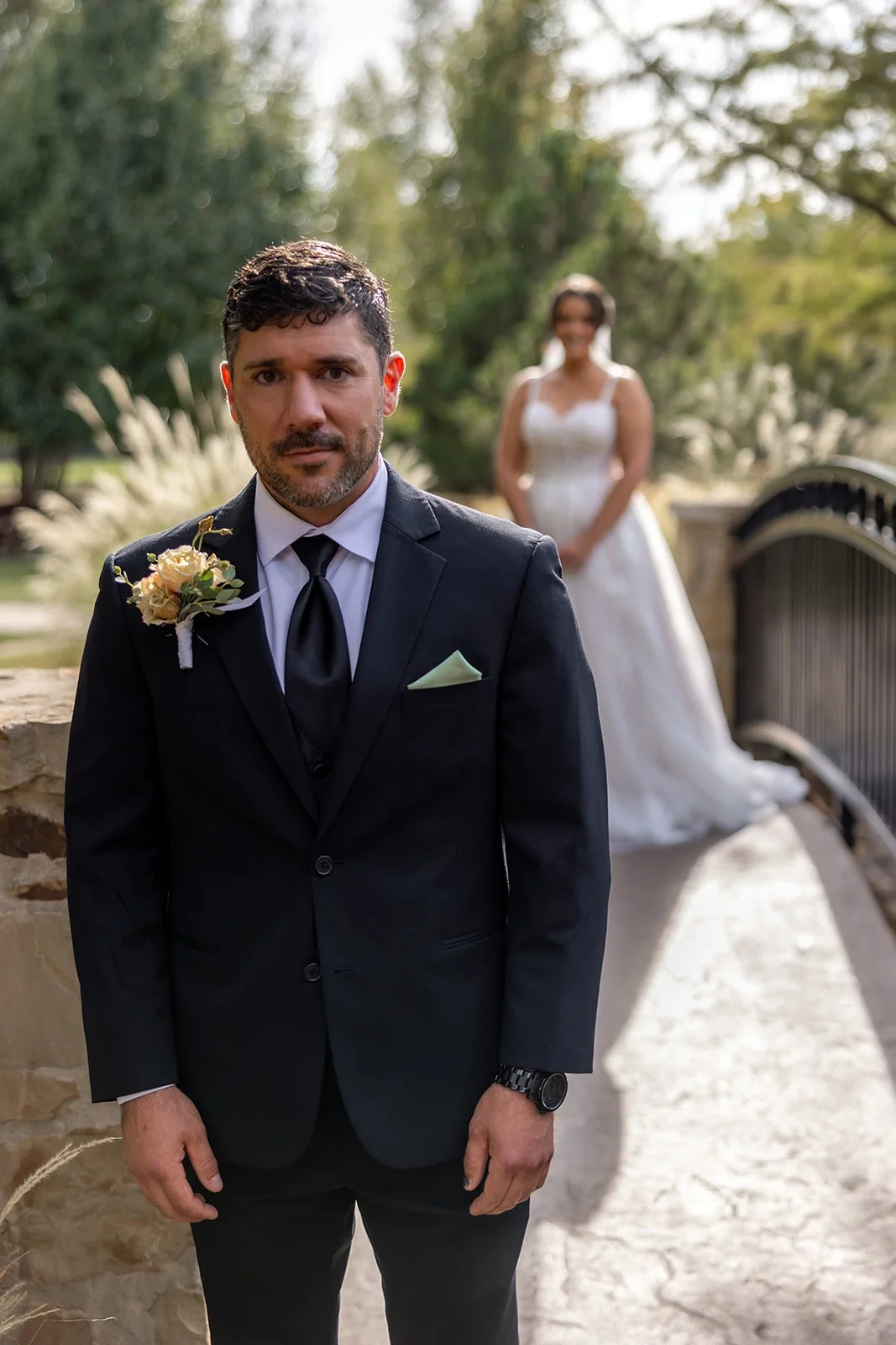 A groom in a black suit with a boutonniere stands in the foreground, with a bride in a white wedding dress in the background, outdoors in a park setting.
