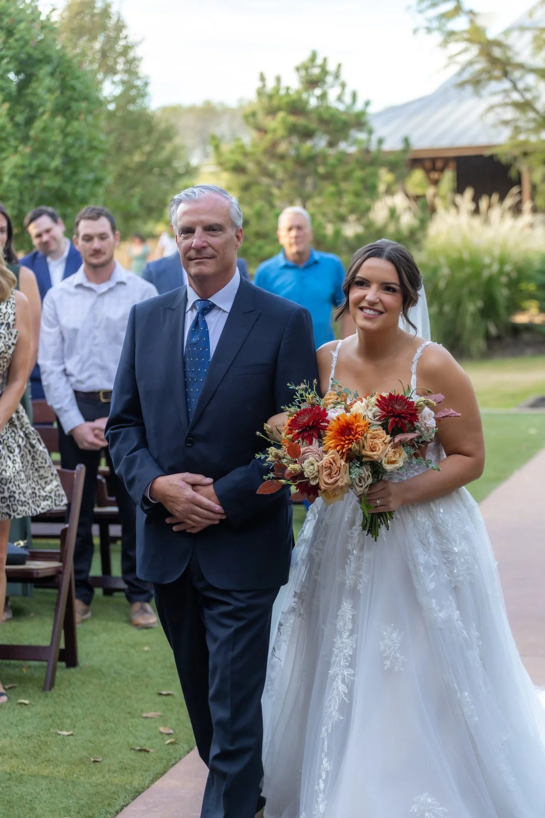 A bride in a white wedding dress holding a bouquet of colorful flowers walking down an outdoor aisle escorted by an older man in a navy suit. Guests are standing behind, outdoors with greenery and a wooden structure in the background.