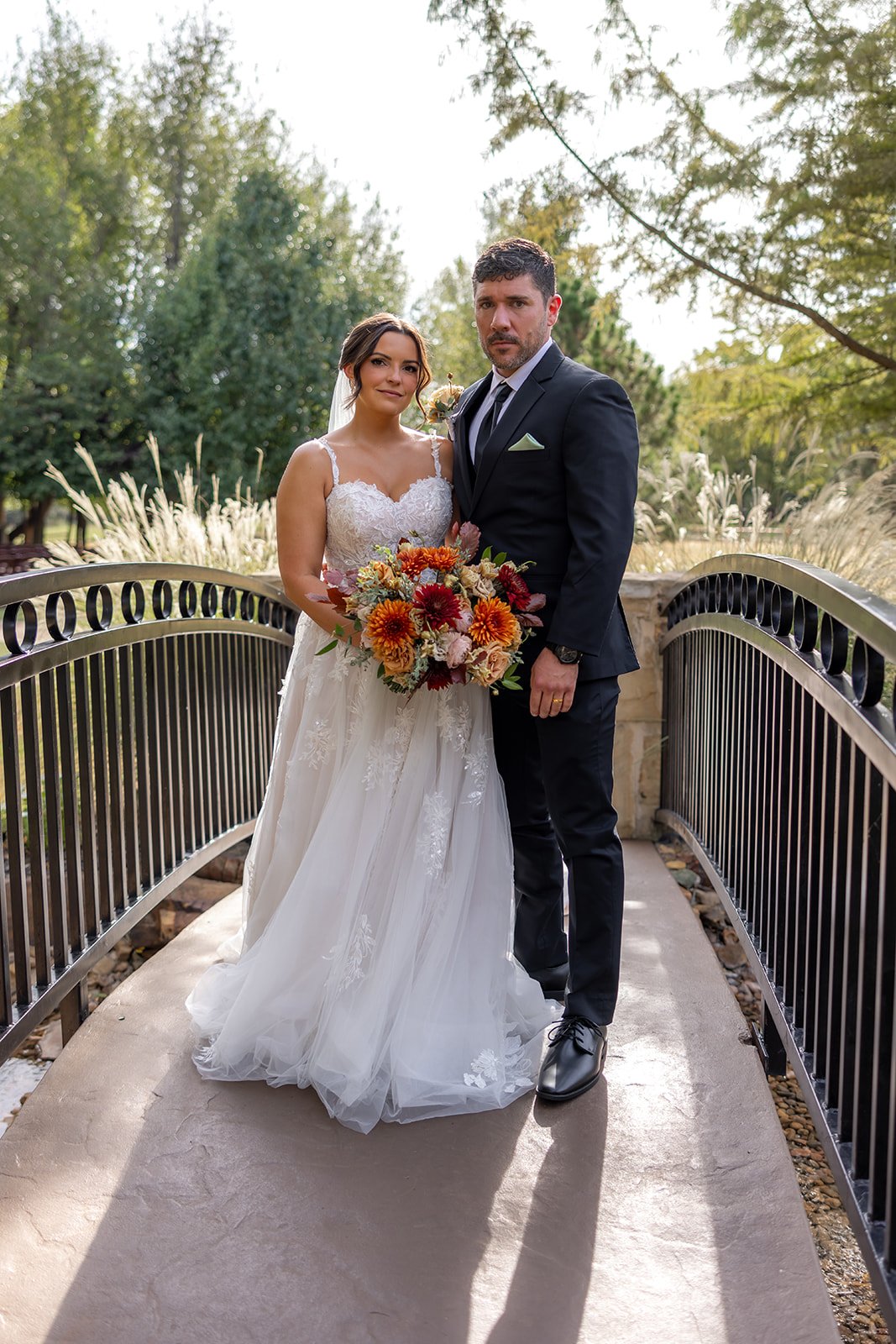 Bride and groom standing on a small bridge outdoors, the bride holding a bouquet of orange, red, and pink flowers, both dressed in wedding attire with trees in the background.
