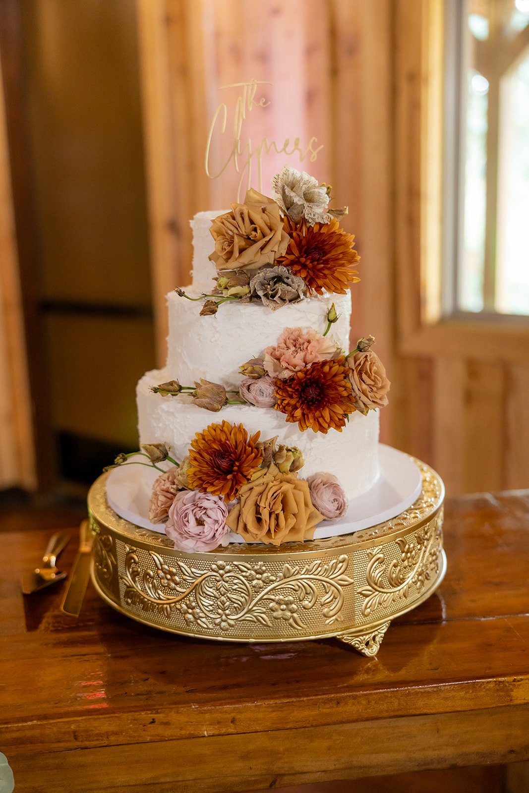 A two-tiered wedding cake decorated with orange, pink, and beige flowers, placed on a decorative gold cake stand on a wooden table.