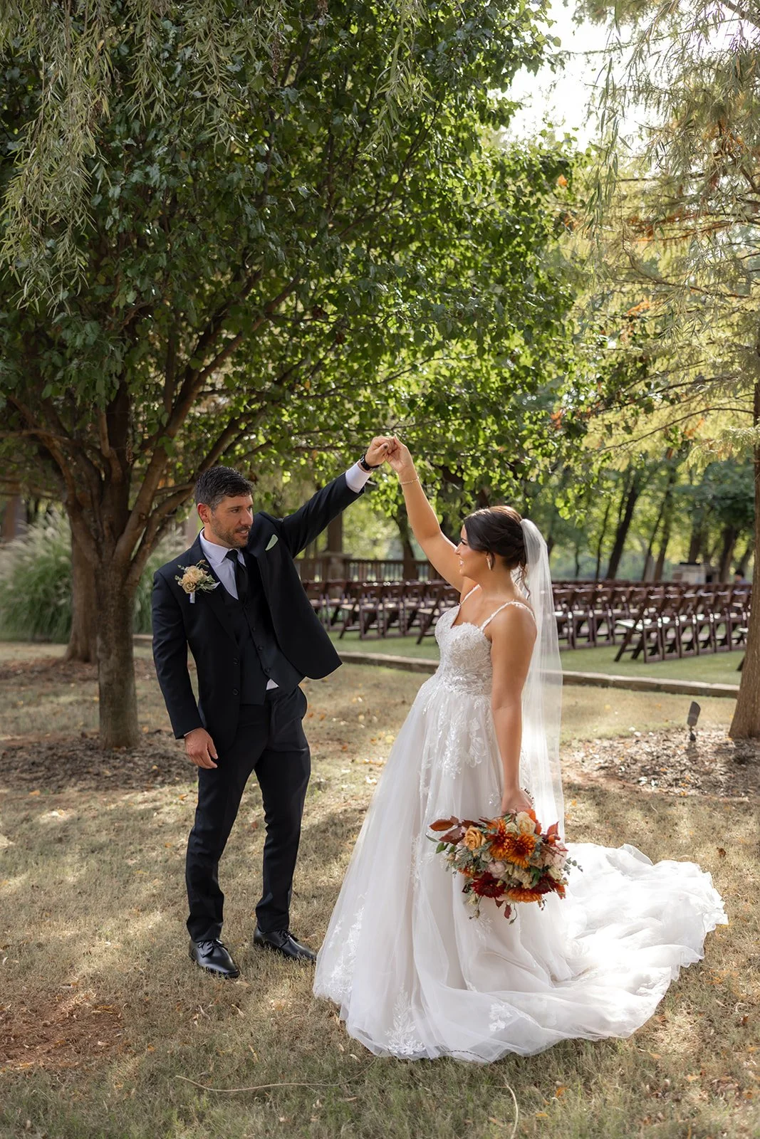 A newlywed couple dancing outdoors; the bride wears a white wedding dress and holds a bouquet, while the groom in a black suit holds her hand as they dance under a large leafy tree in a garden or park setting.