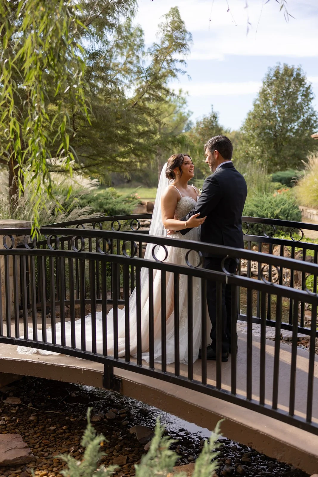 A bride and groom standing on a small bridge outdoors, smiling and looking at each other. The bride is wearing a white wedding dress with lace details and a veil, while the groom is dressed in a black suit. They are embracing each other, surrounded b