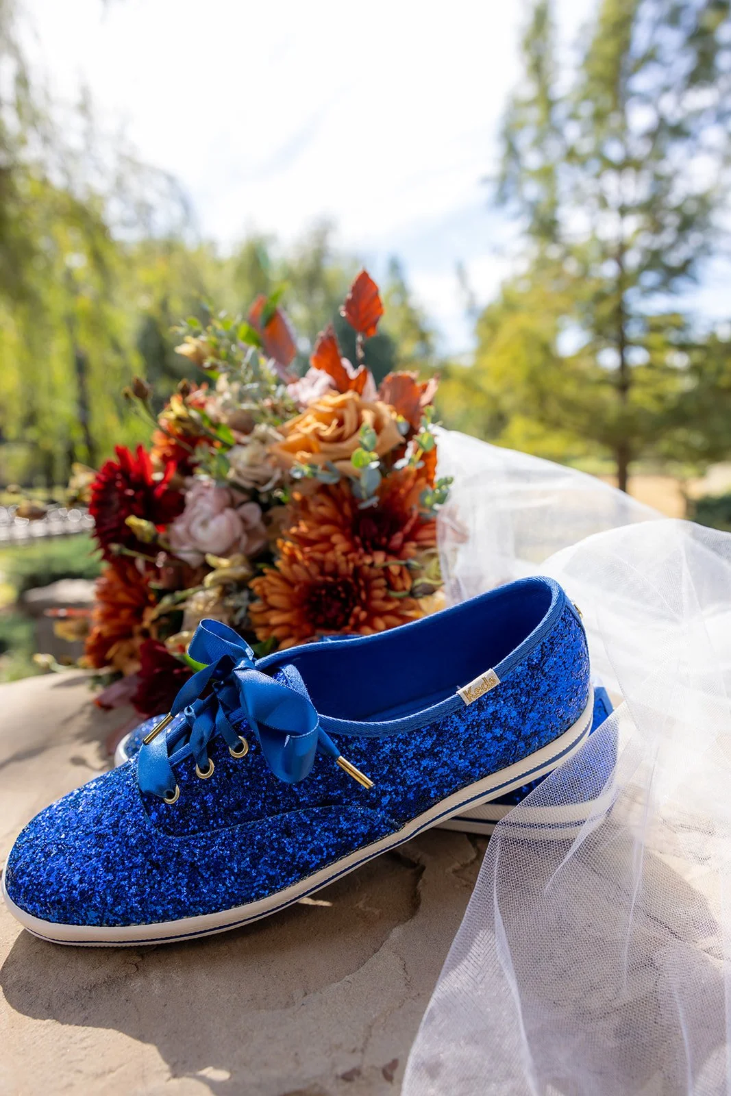 A glittery blue sneaker with satin laces on a rock, with a bouquet of orange, pink, and red flowers behind it, outside in a green, wooded area.