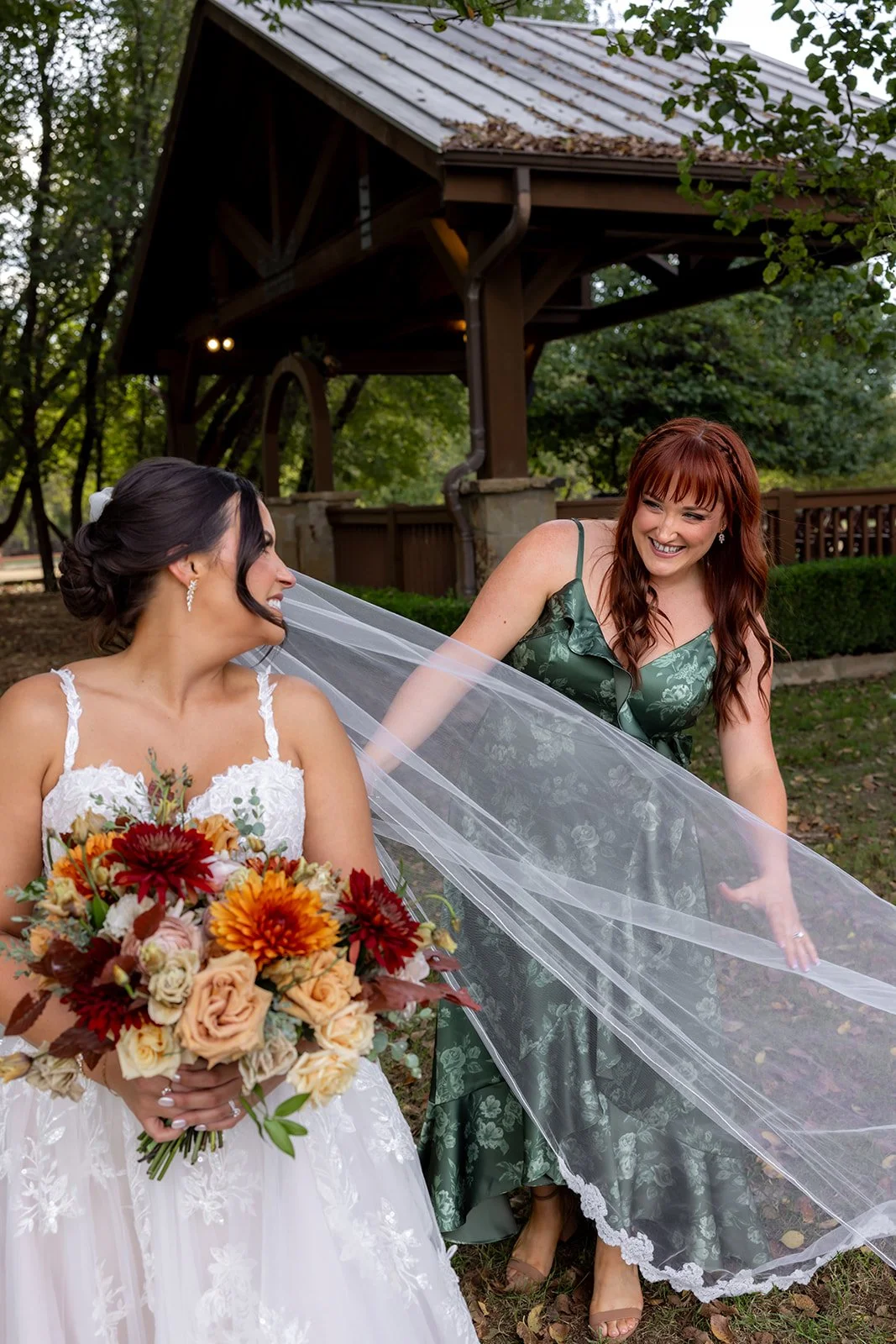 Two women, one in a wedding dress holding a bouquet of flowers and the other in a green dress, laughing outdoors near a gazebo.