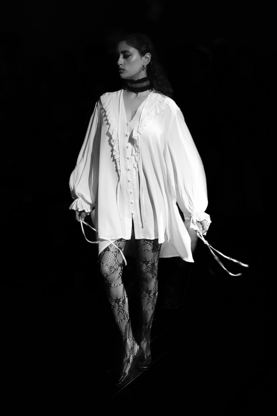 Model walking on runway wearing a loose white blouse with thigh-high lace stockings, under dim lighting.