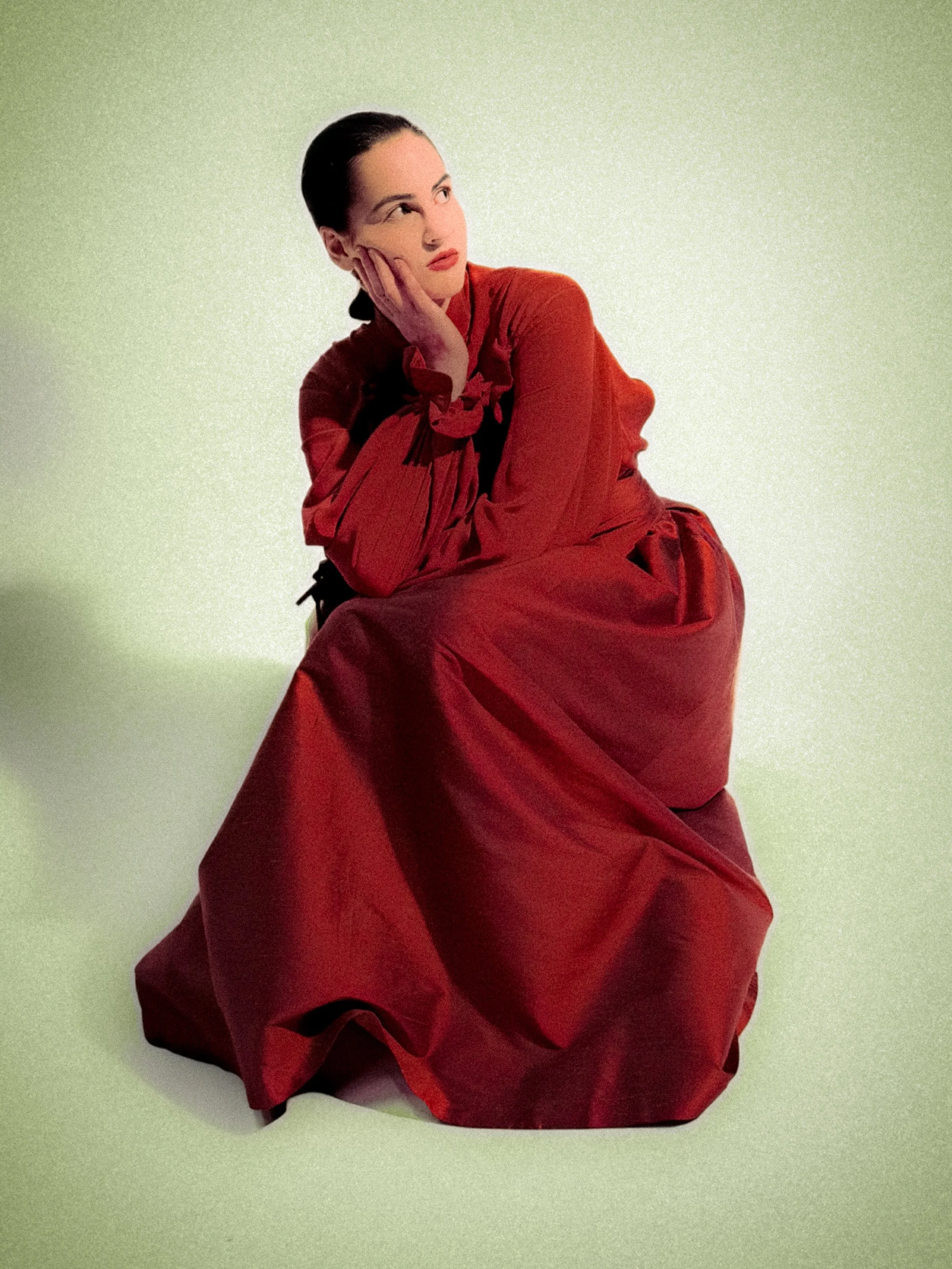 A woman in a long red dress sitting on the floor with her chin resting on her hand, looking thoughtful.