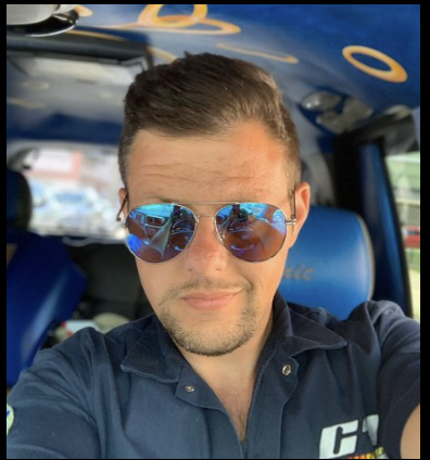 A man wearing blue reflective sunglasses and a dark blue button-up shirt taking a selfie inside a vehicle.