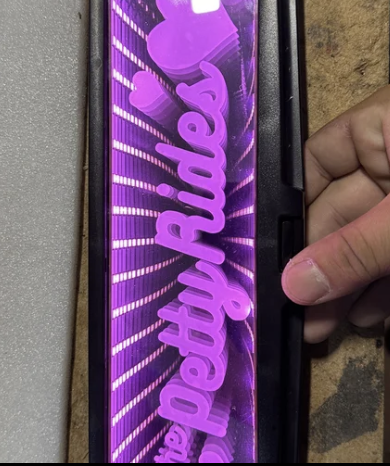 A close-up of a pink neon sign that reads 'the petty rides' with heart shapes, held by a person's hand against a black surface on a wooden table.