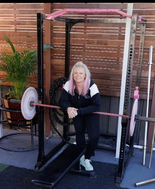 A woman with long hair and a smile sitting on a weightlifting bench inside a home gym, holding a barbell with pink weights. The gym has wooden fencing and some potted plants in the background.