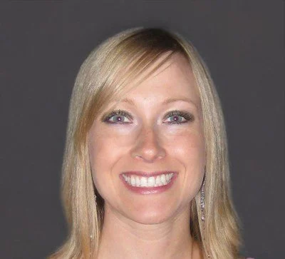 Close-up of a smiling woman with blonde hair, wearing earrings and makeup against a gray background.