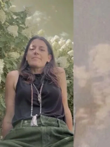 A woman with long brown hair wearing a black sleeveless top and green pants, standing outdoors with trees and cloudy sky in the background.