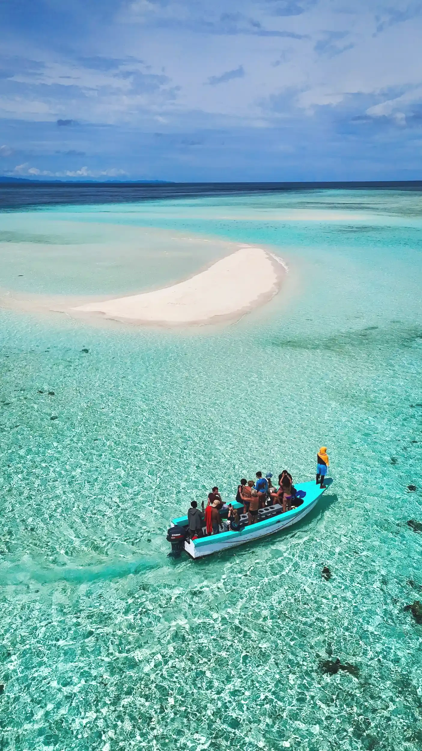 Boat with people sailing on clear turquoise water near a white sandbar and a distant shoreline under a partly cloudy blue sky.