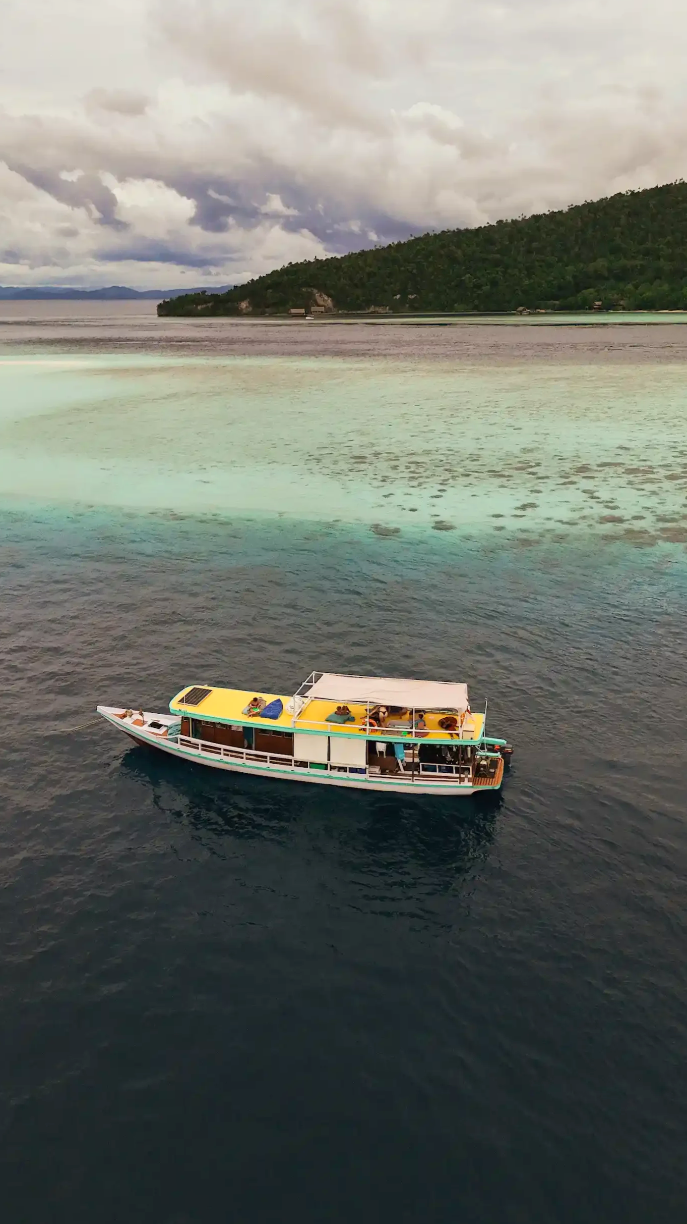 A boat floating on dark water with a green shoreline and forested hill in the background on a cloudy day.