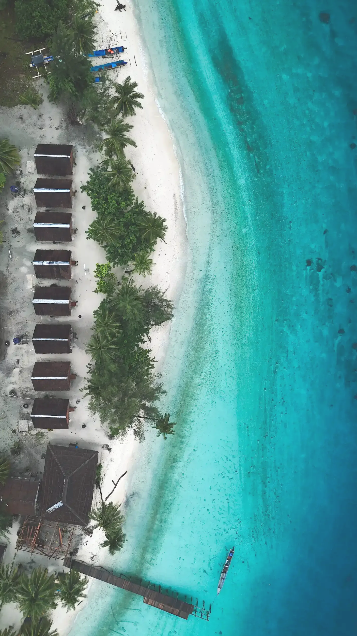 Aerial view of a tropical beach with white sand, clear blue water, palm trees, beach huts, and a dock with a boat