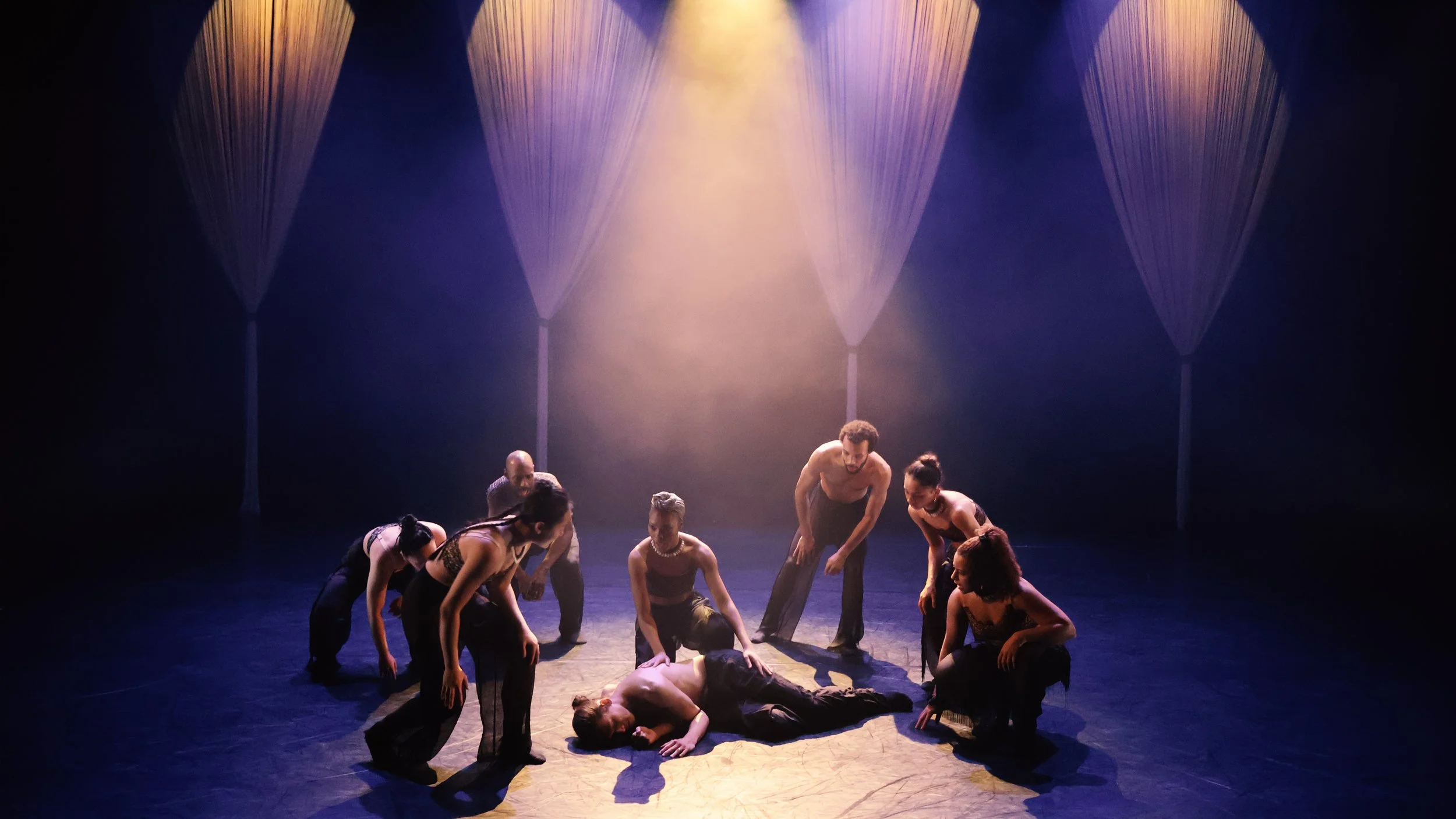 Ensemble gathered under warm amber and violet stage lighting with suspended string installations in Tellus by Dickson Mbi, stage design by London set designer Ruby Law.