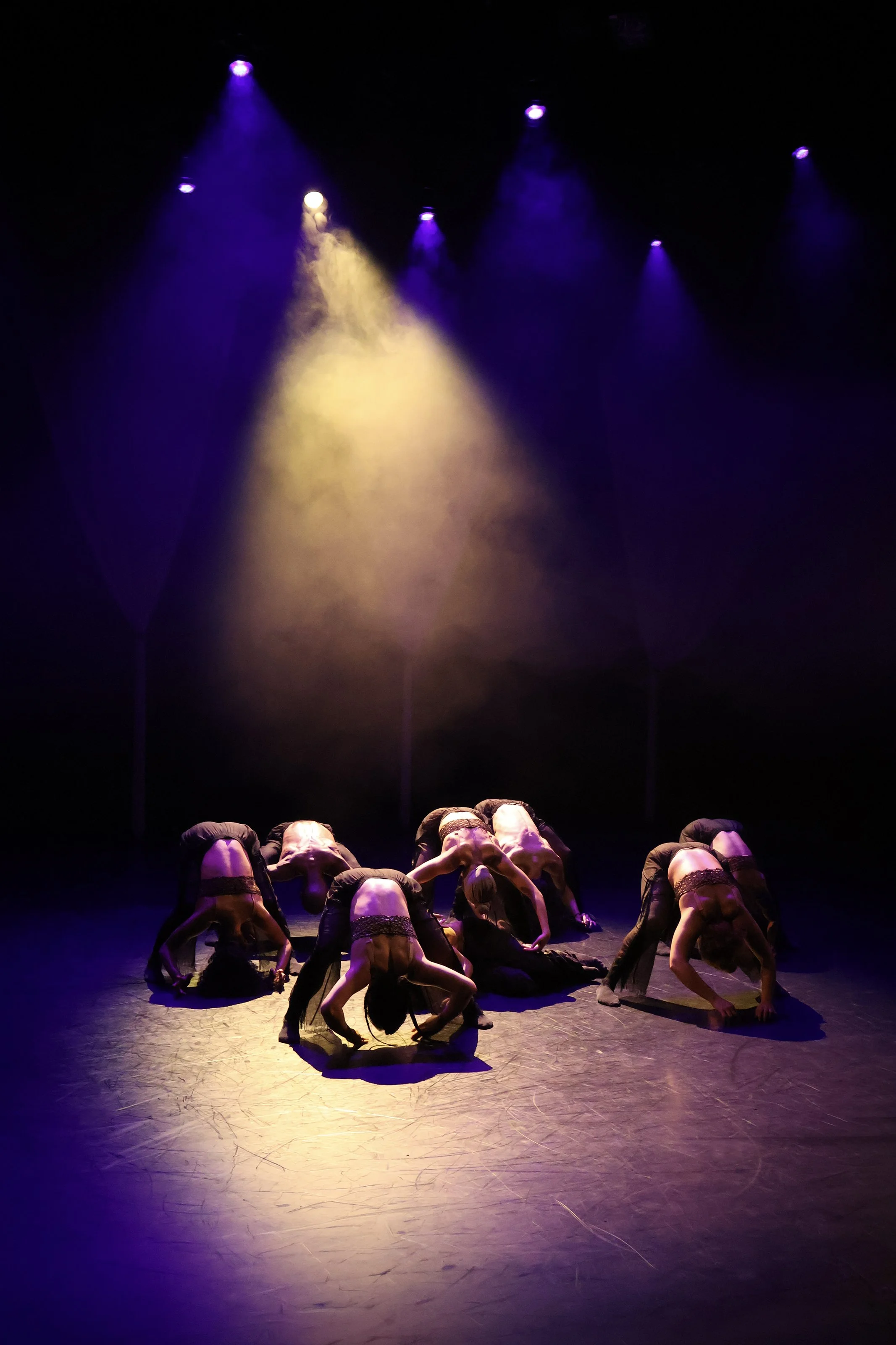 Contemporary dancers arching in unison beneath purple spotlights and haze in Tellus by Dickson Mbi, theatrical lighting and set design by London designer Ruby Law.