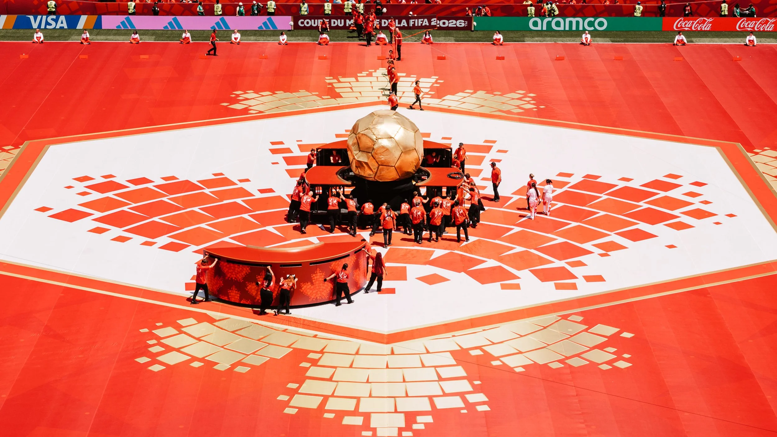Wide stadium view of the opening ceremony stage, featuring the central Trionda Ball surrounded by the Canadian mosaic graphics across the performance floor.