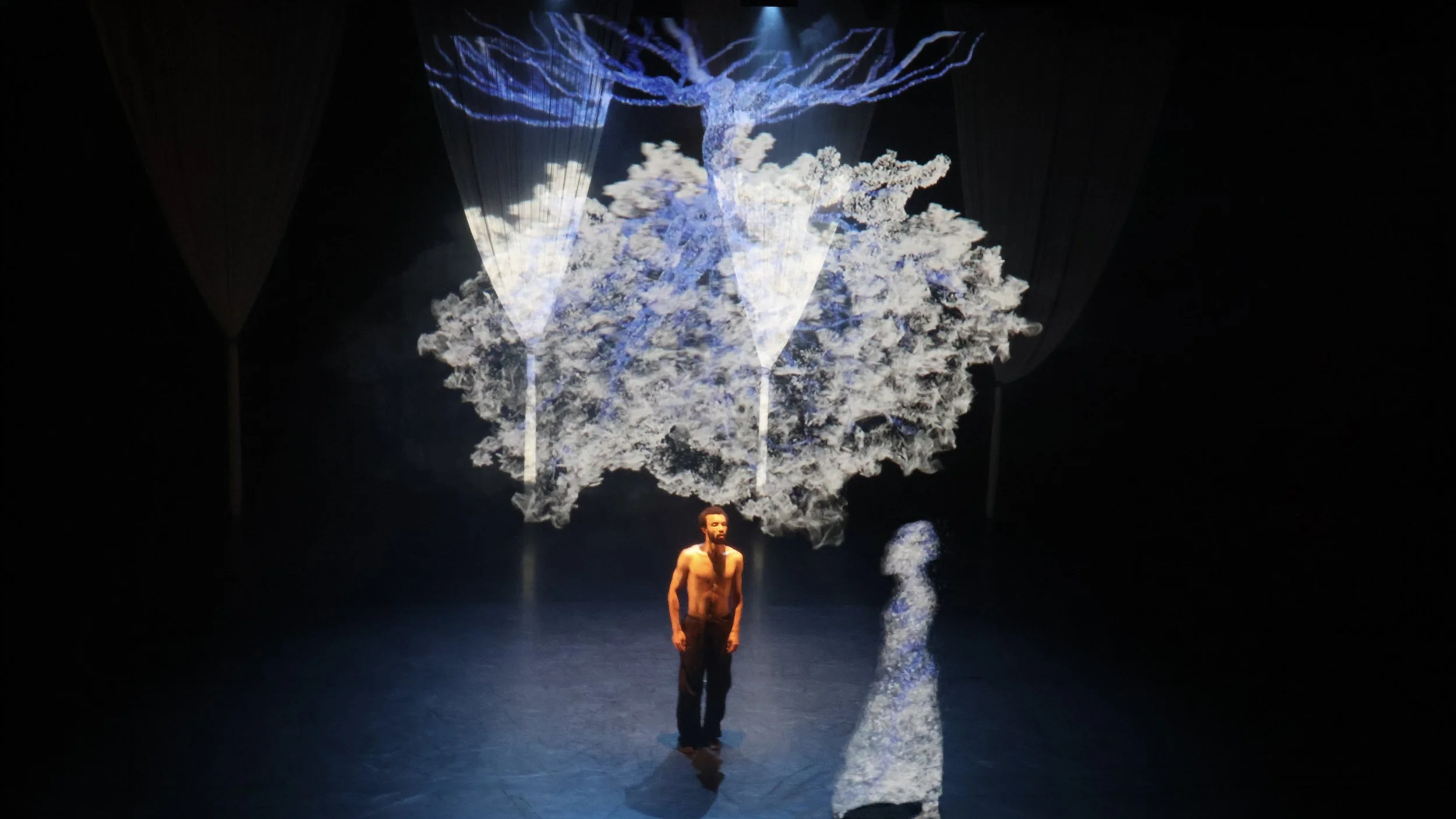 Solo performer facing projected ghostlike figure beneath digital tree canopy in Tellus by Dickson Mbi, contemporary stage design by London set designer Ruby Law.