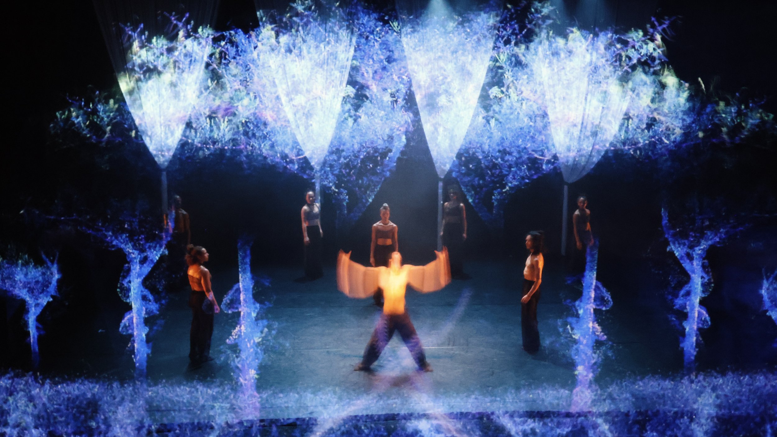 Projection mapping forming luminous blue tree structures around central dancer in Tellus by Dickson Mbi, immersive stage design by London set designer Ruby Law.
